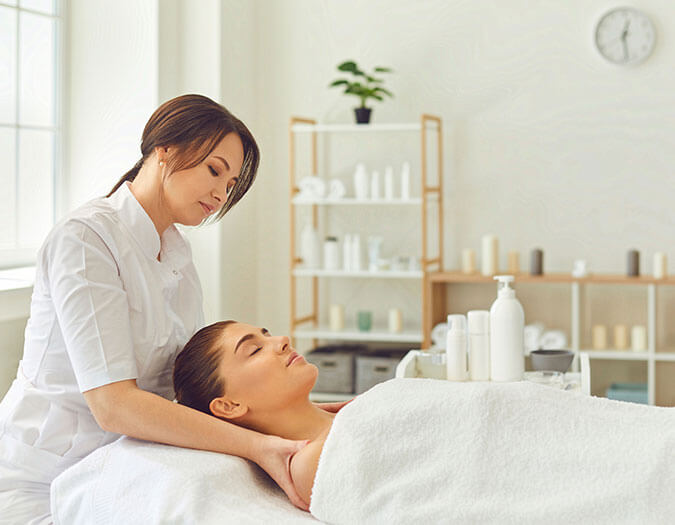 Young woman lying on bed with eyes closed enjoying a face and neck massage done by a professional therapist