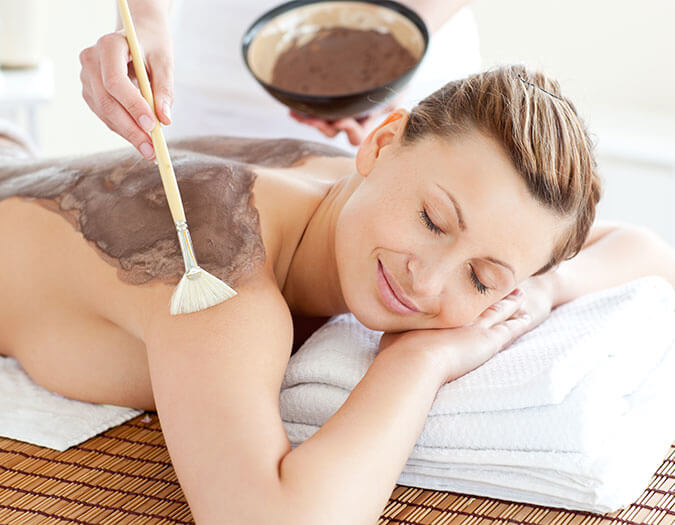 Woman laying down on bed, getting a body mud wrap by a therapist